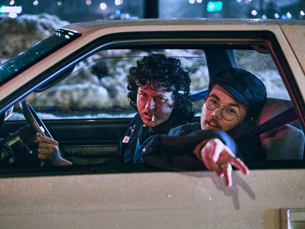 Two people in a car at night, one smoking and the other pointing, with a snowy street outside.