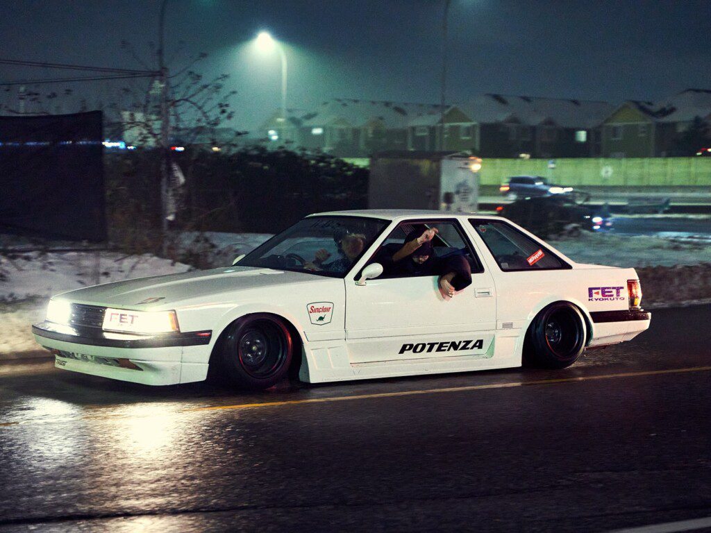 White sports car with Potenza decals driving at night, passenger gesturing out the window, wet road reflecting lights.