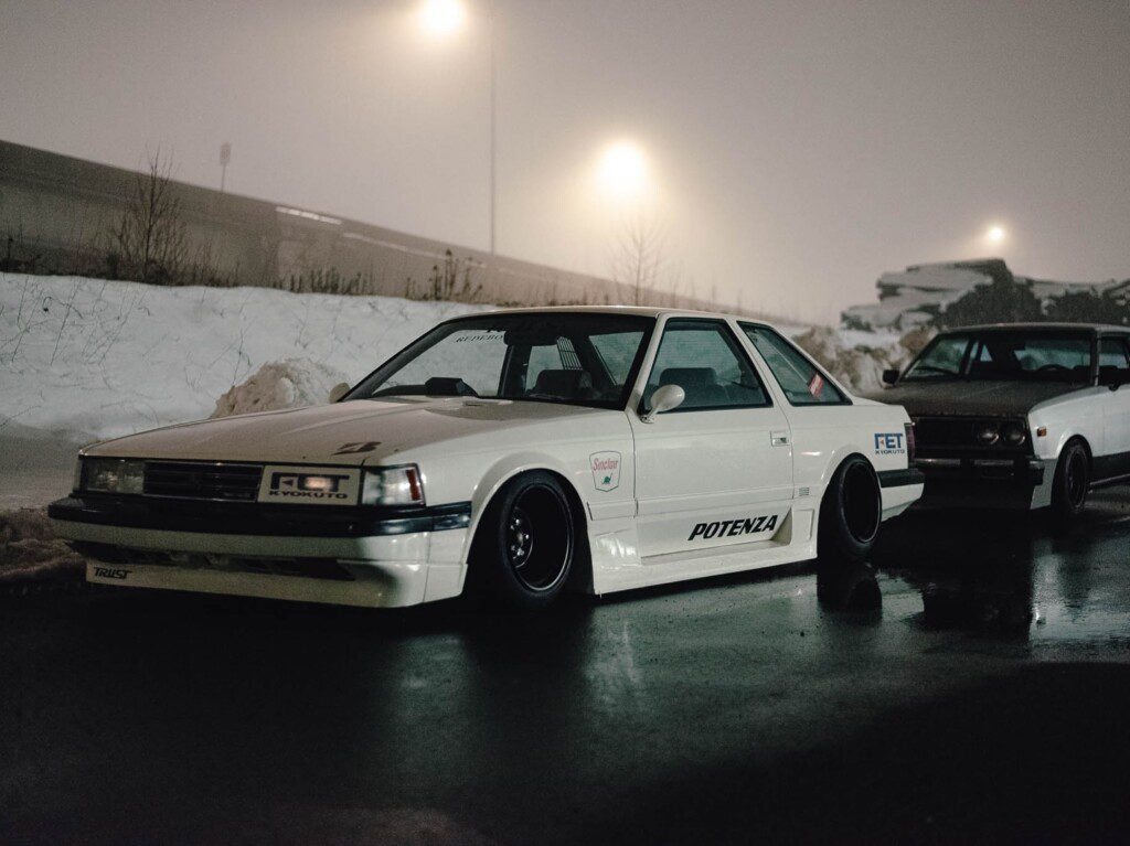 Modified white vintage car on wet pavement, parked at night with snow and foggy streetlights.