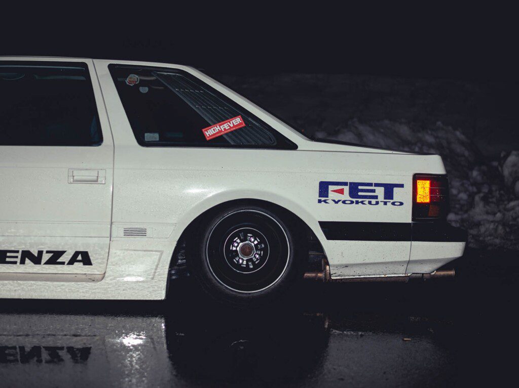 Vintage white car with FET Kyokuto decal parked on wet pavement at night. Classic design with striking taillight.