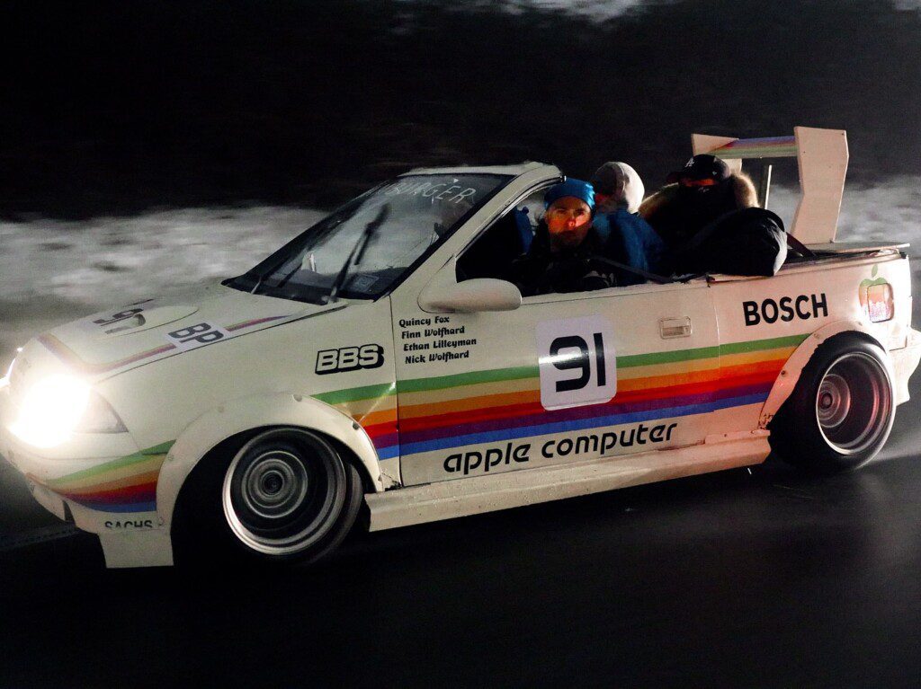 Retro car with Apple Computer branding and rainbow stripes on a snowy road, filled with passengers wearing winter gear.