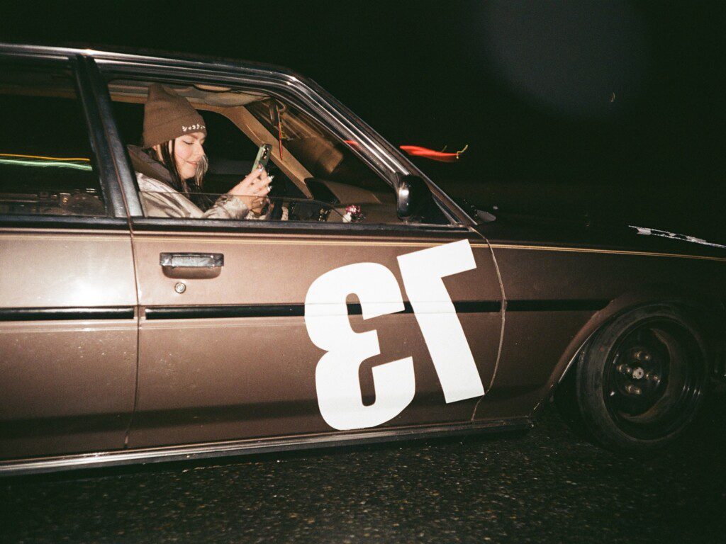Person in beanie using phone inside a parked vintage car with number 13 on the side, at night.