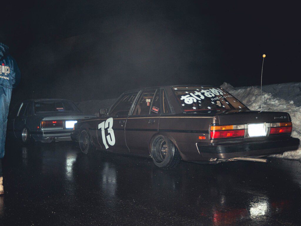 Vintage cars parked on wet road at night, featuring racing decals and Japanese text on rear windshield.