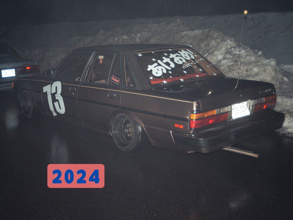 Retro car with number 73 parked on wet road, snow in background, Japanese text on window, 2024 in colorful overlay.