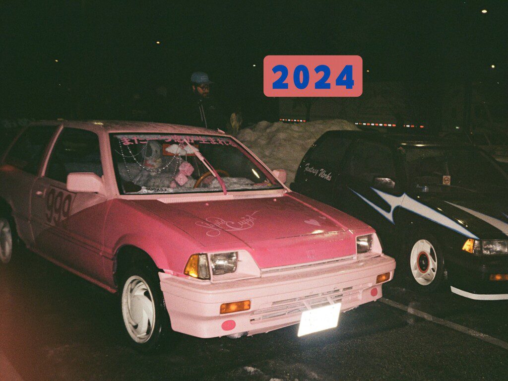 Pink retro car with decals, number 999, parked next to a black car at night. Sign reading 2024 above.