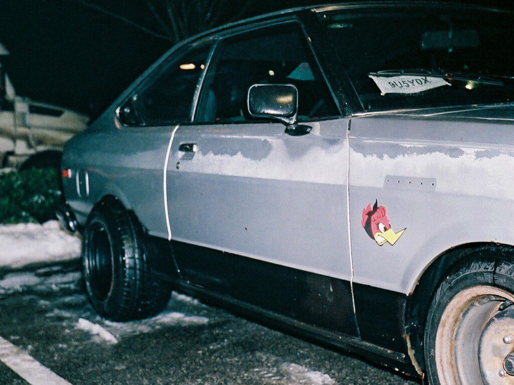 Vintage gray car with black rims, parked in snow, featuring a red cartoon bird decal on the side.