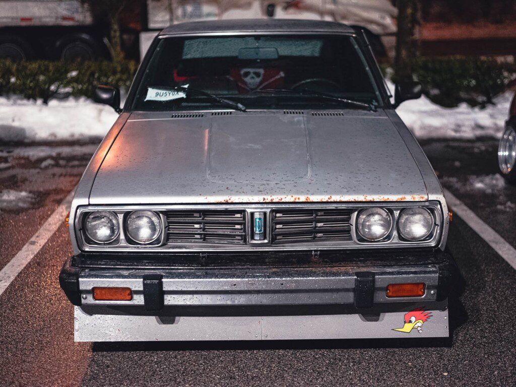 Vintage silver car with distinctive rectangular headlights and a cartoon bird sticker, parked in a snowy lot.