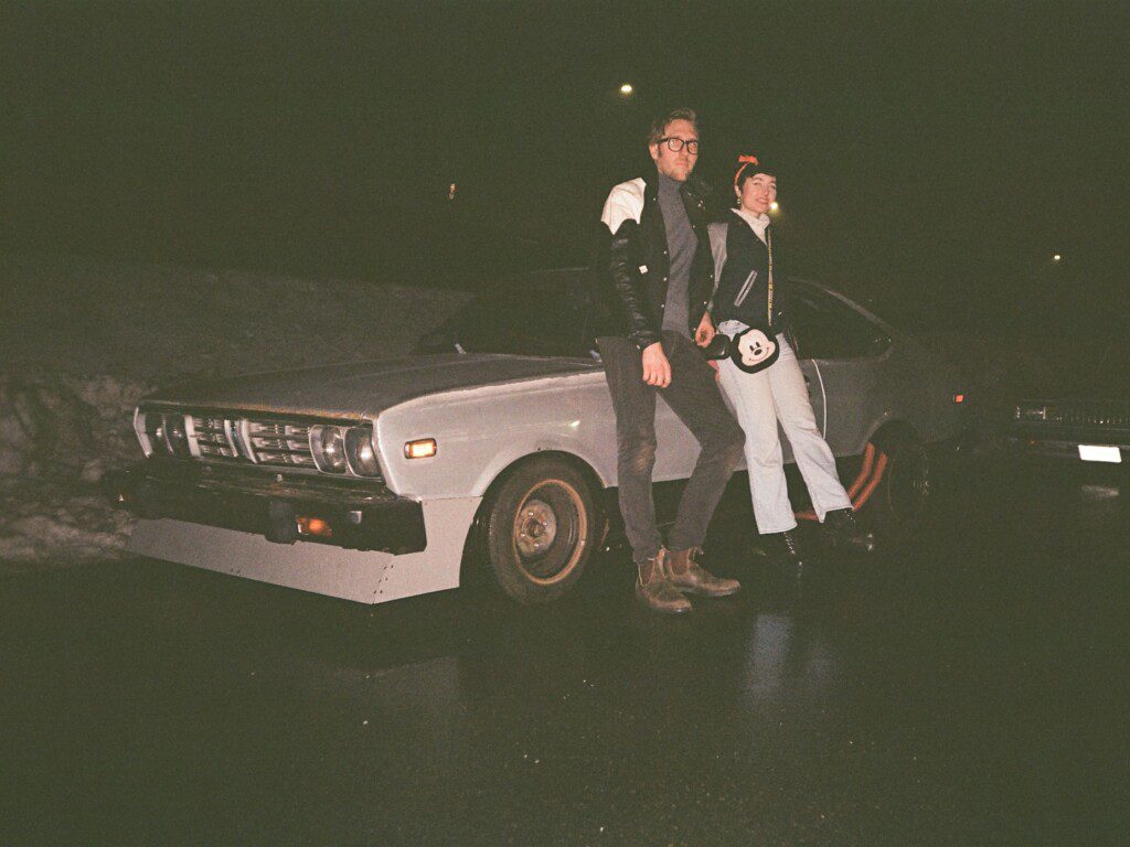 Two people leaning on a vintage car at night, wearing retro attire, on a dimly lit street.