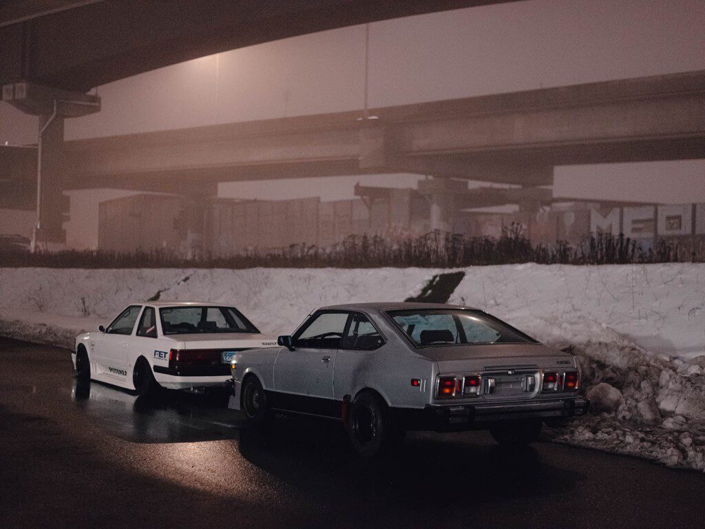 Vintage cars parked under misty highway, near snowy road. Retro night scene with classic vehicles.