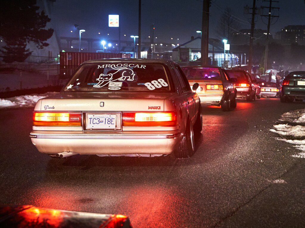 Cars queued at night on a wet road, showcasing classic sedans with custom decals and headlights illuminating the street.