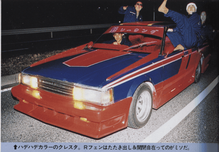 Customized vintage car with colorful paint job and people posing on a nighttime street.