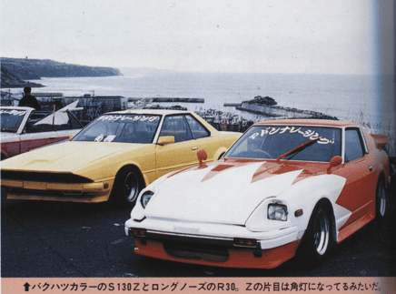Vintage S130Z and R30 cars with custom decals parked near the ocean with a scenic view in the background.