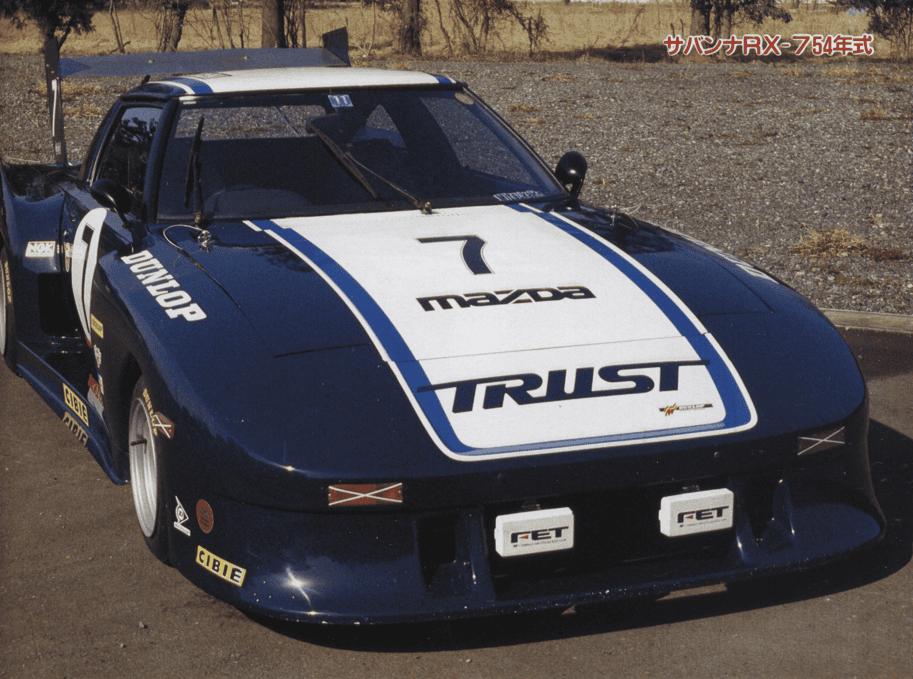 Vintage Mazda race car with Trust and Dunlop branding, parked on a sunny day.