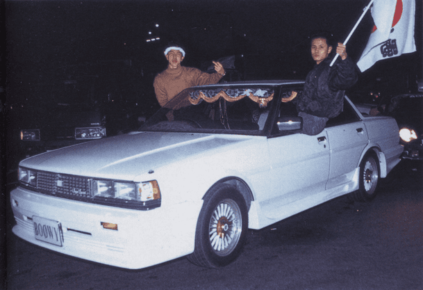 Vintage white car at night with two people, one waving a flag; retro street scene.