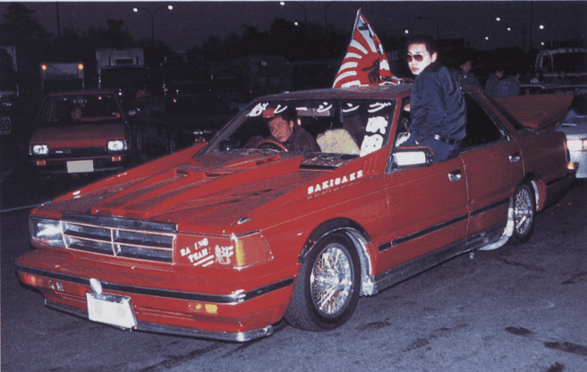 Customized red car with Japanese text and flag, parked at night with a person sitting on the hood.