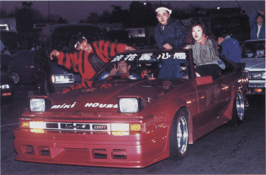 Classic red sports car with passengers at night, showcasing 80s style and custom modifications.