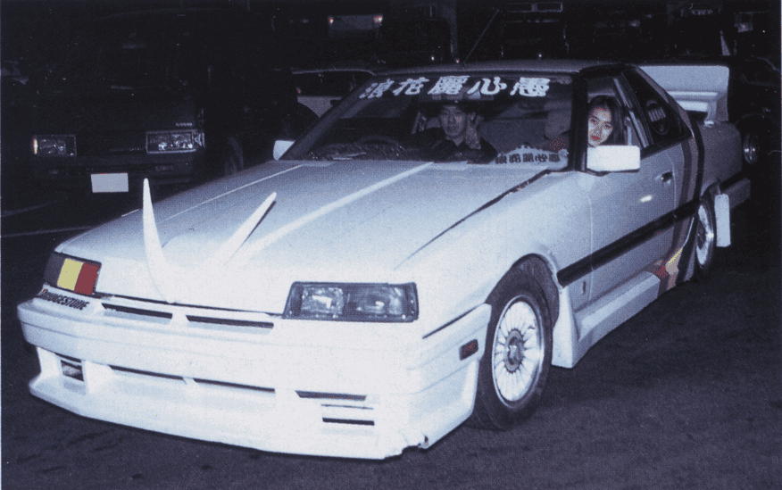 Customized white car with bold decals and unique front adornments, parked with passengers inside at night.