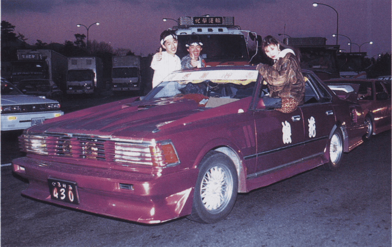 Three people enjoying a night out with a custom red car in a parking lot, surrounded by other vehicles.