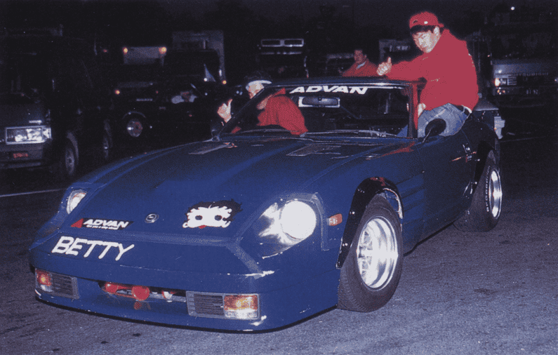 Blue vintage car with Betty decal at a nighttime event, driver wearing a red jacket and cap, parked among vehicles.