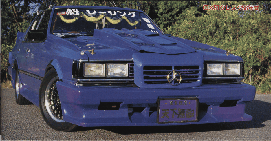 Blue vintage car with modified body kit and front grille, parked on a road with trees in the background.