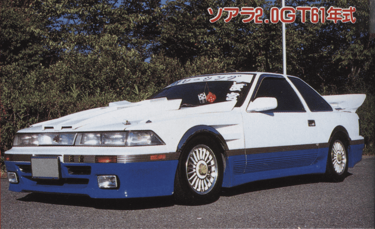 Vintage white and blue modified car with decals, parked outdoors with greenery in the background.