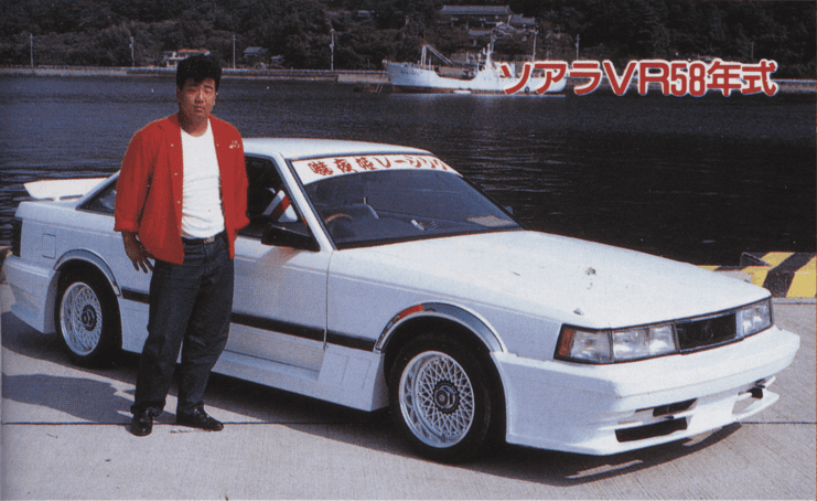 Man standing beside a classic white Toyota Soarer VR with scenic waterfront backdrop.