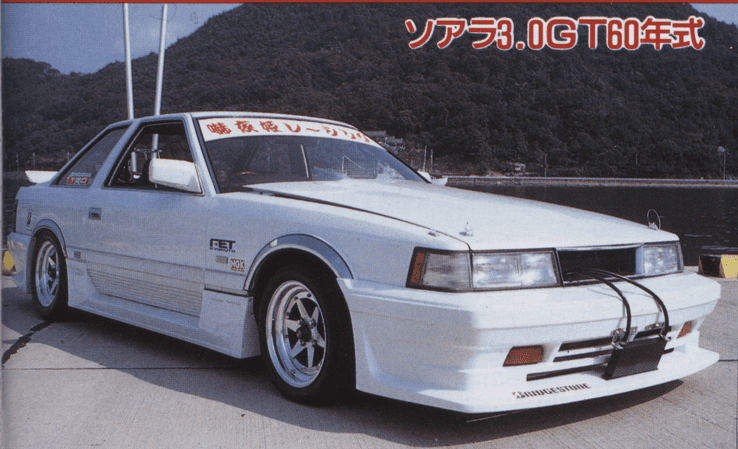 Vintage white sports car parked by a waterfront with bold decals, showcasing classic 80s Japanese automotive design.