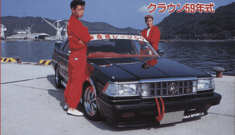 Vintage Toyota Crown car with two people in red jumpsuits by the waterfront, Japanese text overhead.