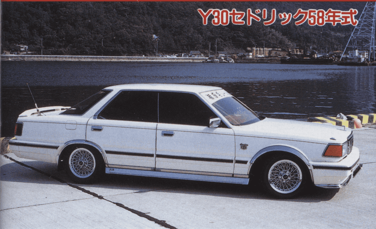 Vintage white sedan parked by the waterfront, showcasing stylish rims and a classic design in a serene setting.
