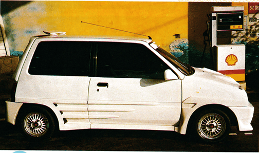 White compact car with sporty body kit parked at a Shell gas station with vibrant mural background.