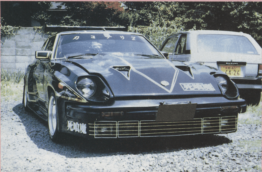 Customized vintage car with unique decals and black paint parked beside a white vehicle outdoors.