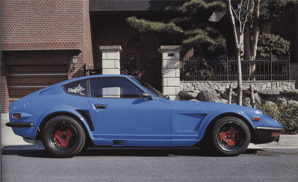 Vintage blue sports car parked in front of a brick house, showcasing classic design and vibrant red wheels.
