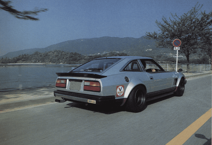 Vintage sports car cruising by a lake with mountains in the background on a sunny day.