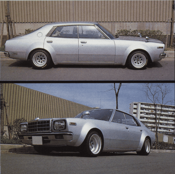 Classic vintage sedan with silver body and sporty wheels, parked on urban street with buildings in the background.
