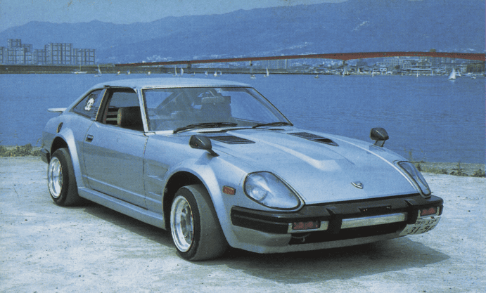 Classic silver sports car parked by a serene waterfront, with a scenic bridge in the background under a clear sky.
