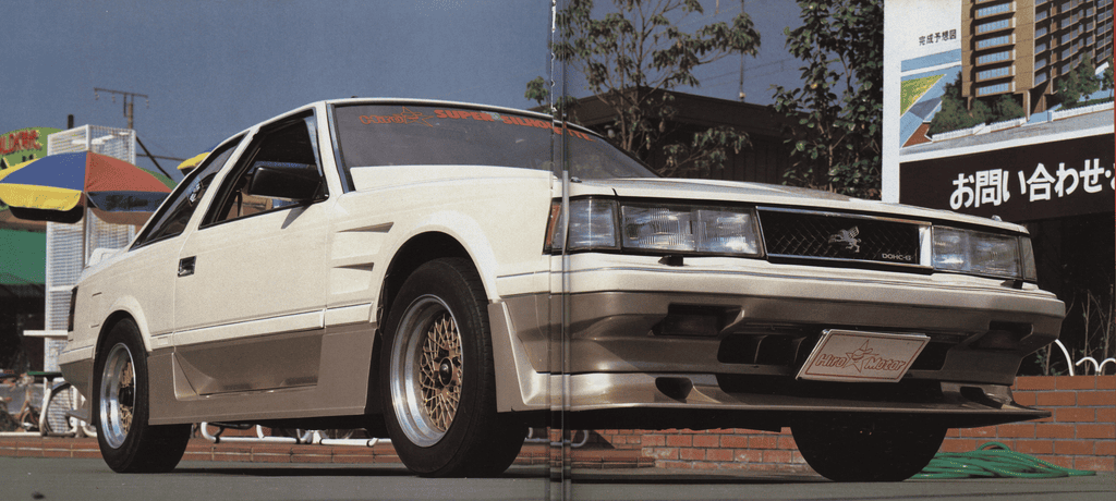 Vintage white sports car with gold rims parked outdoors on a sunny day, showcasing classic 1980s design.
