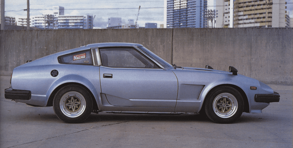 Vintage silver sports car parked in urban setting, sleek design with retro styling and gold alloy wheels.