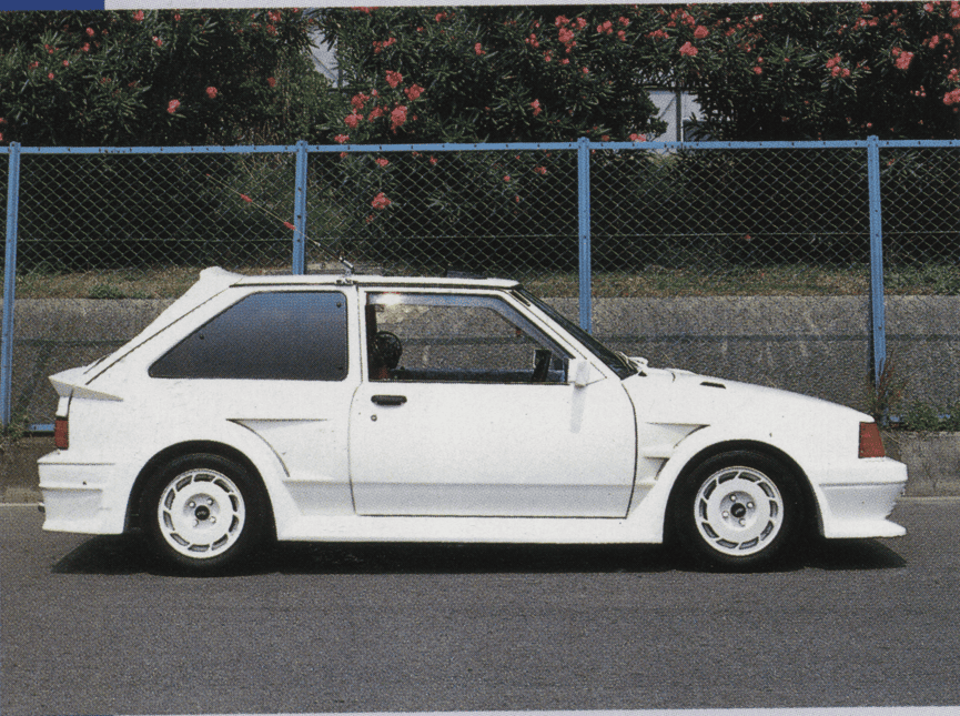 White classic hatchback with sporty body kit parked in front of blue fence and green foliage background.