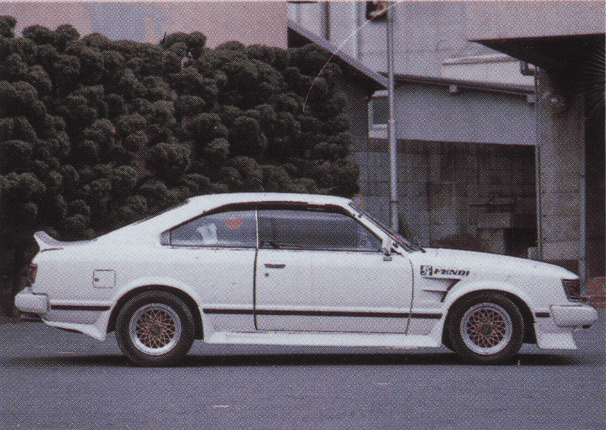 Vintage white sports car with custom wheels parked on a city street, surrounded by greenery.