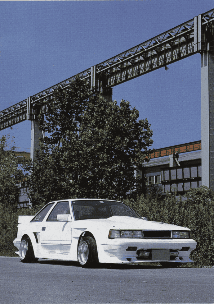 A vintage white sports car parked, industrial background with steel beams and clear blue sky.