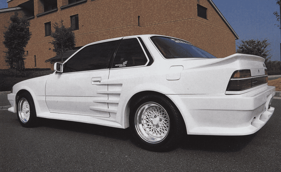 White vintage sports car with custom body kit and mesh wheels, parked near a brick building.